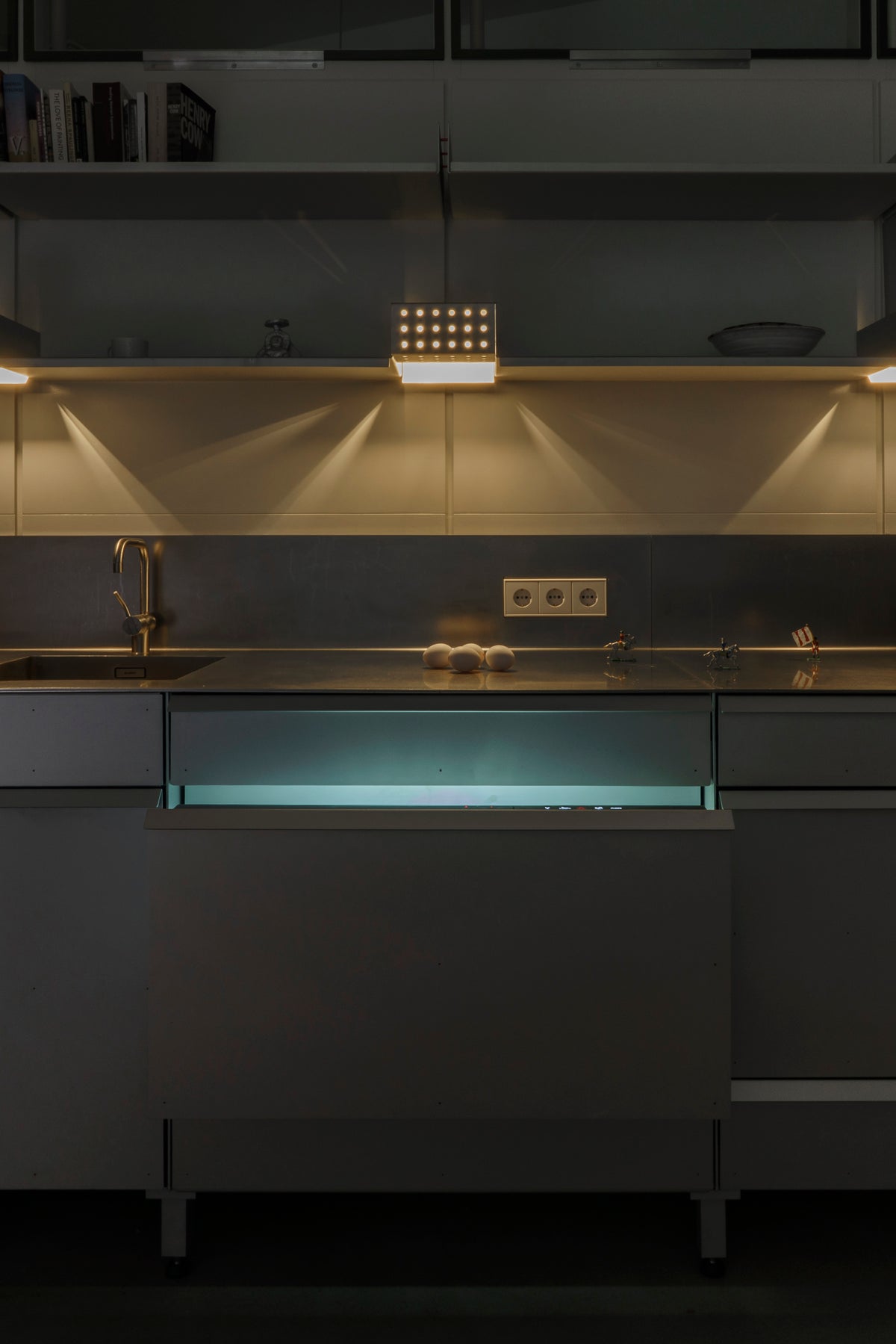 The kitchen at night, a fridge drawer glowing turquoise beneath the stainless-steel worktop