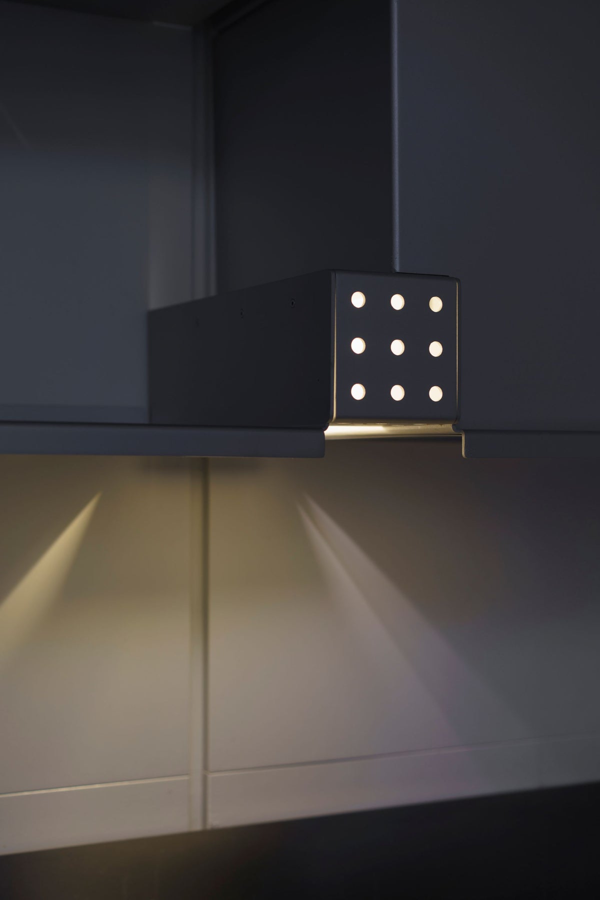 Nine-hole bespoke shelf light mounted under a cabinet, throwing overlapping beams down the panel below